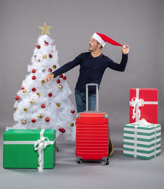 Front view young man holding his santa hat standing near white xmas tree and presents on grey isolated