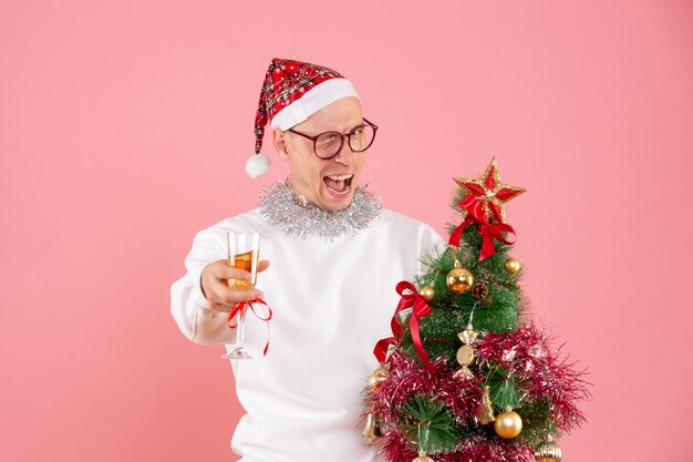 Front view of young man holding little christmas tree and drink on pink wall