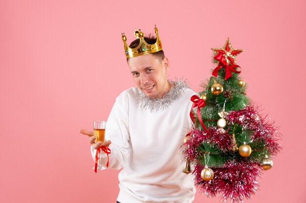 ピンクの壁に飲み物と小さなクリスマスツリーを保持している若い男の正面図