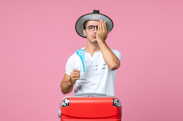 Front view of young man holding plane ticket for vacation on light-pink wall