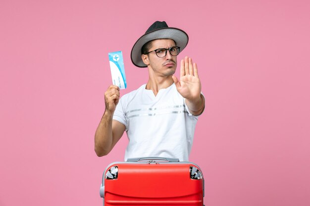 Front view of young man holding plane ticket for vacation on pink wall