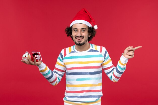 Вид спереди молодой человек держит пластиковые игрушки на красной стене красные человеческие новогодние праздники