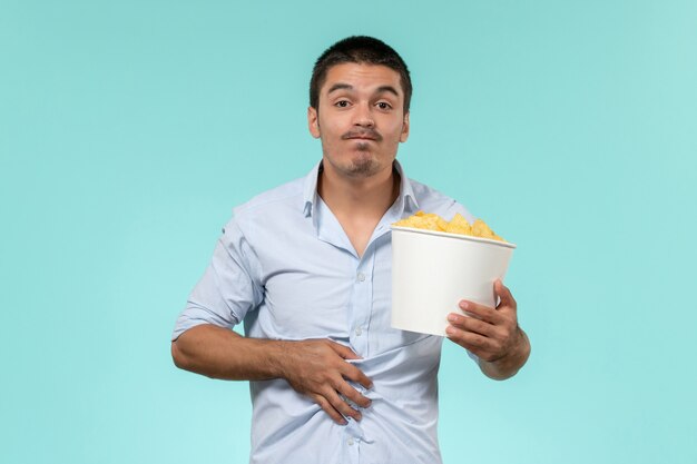 Вид спереди молодой человек, держащий картофельные чипсы на голубой стене, одинокий удаленный мужской кинотеатр