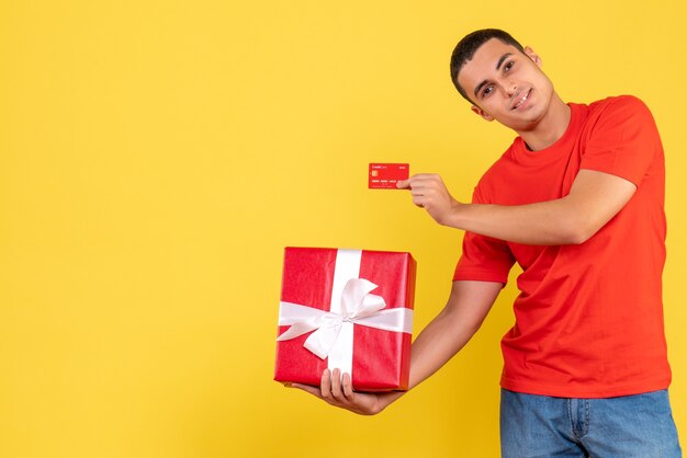 Front view of young man holding present and bank card on the yellow wall