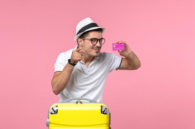Front view of young man holding purple bank card on summer vacation on pink wall