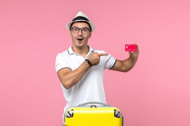 Front view of young man holding red bank card on summer vacation on light pink wall