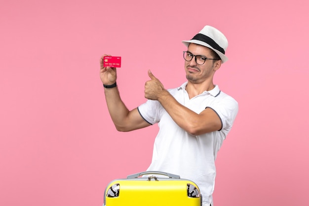 Front view of young man holding red bank card on vacation on a pink wall
