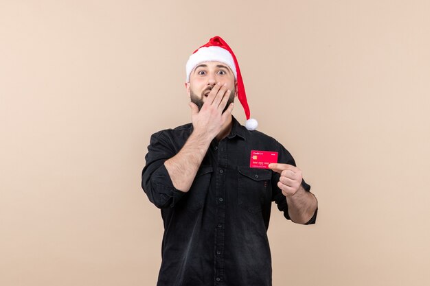 Front view of young man holding red bank card with surprised expression on a pink wall