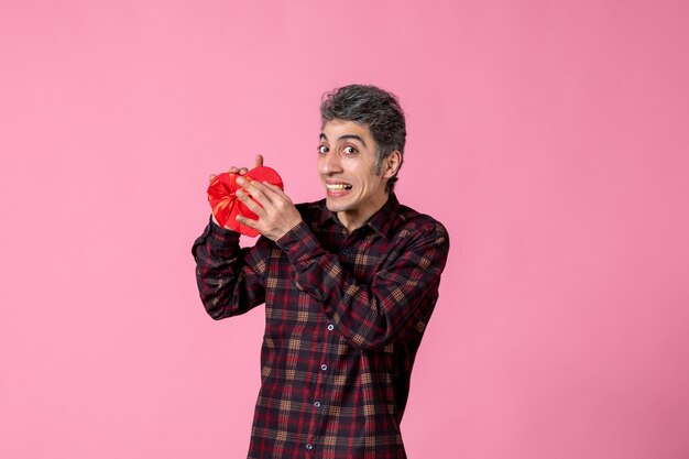 Front view young man holding red heart shaped present on pink wall