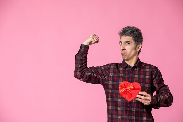 Front view young man holding red heart shaped present on pink wall