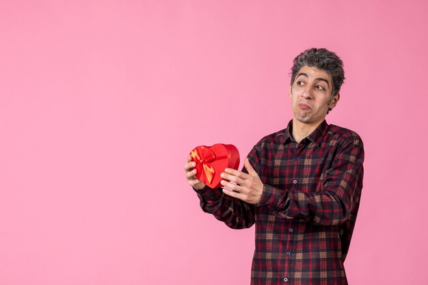 Front view young man holding red heart shaped present on pink wall