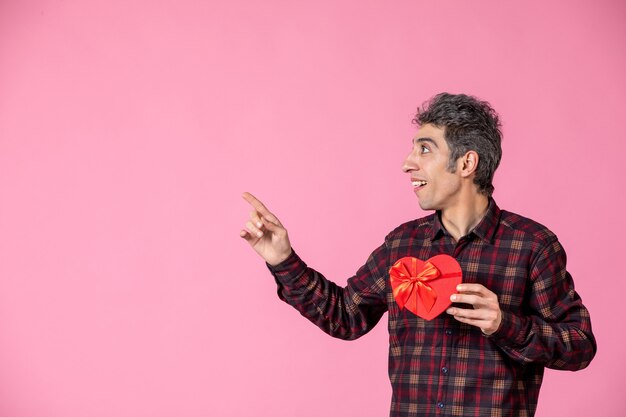 Front view young man holding red heart shaped present on pink wall