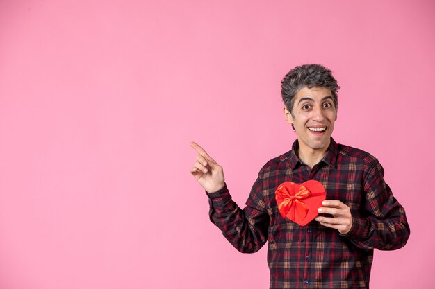 Front view young man holding red heart shaped present on pink wall