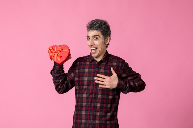 Front view young man holding red heart shaped present on pink wall