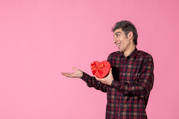 Front view young man holding red heart shaped present on pink wall