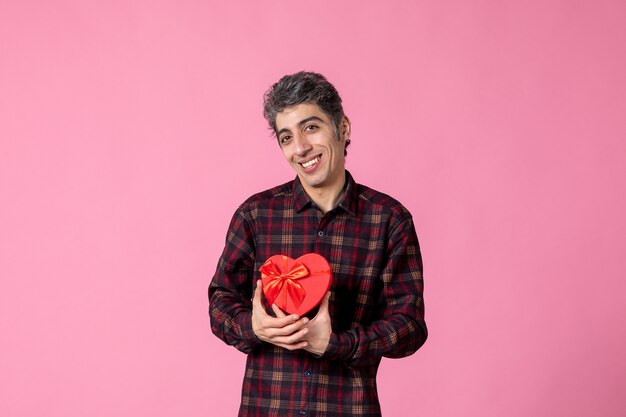 Front view young man holding red heart shaped present on pink wall