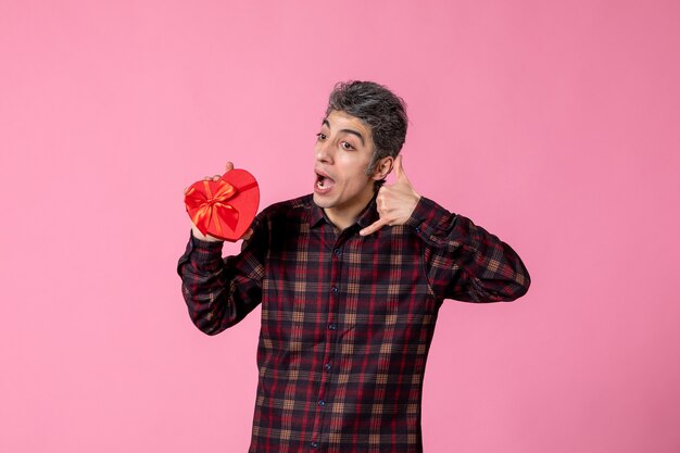 Front view young man holding red heart shaped present on pink wall