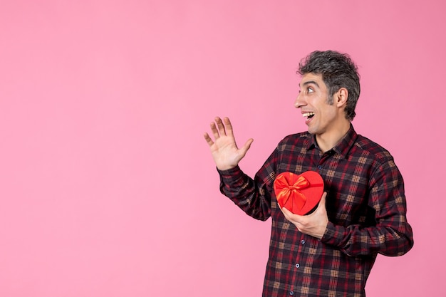 Вид спереди молодой человек, держащий красный подарок в форме сердца на розовой стене