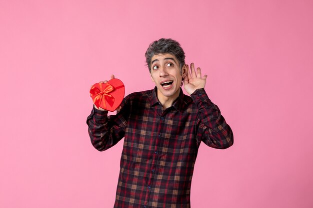 Front view young man holding red heart shaped present on pink wall