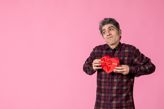 Front view young man holding red heart shaped present on pink wall