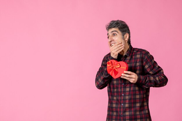 Front view young man holding red heart shaped present on pink wall