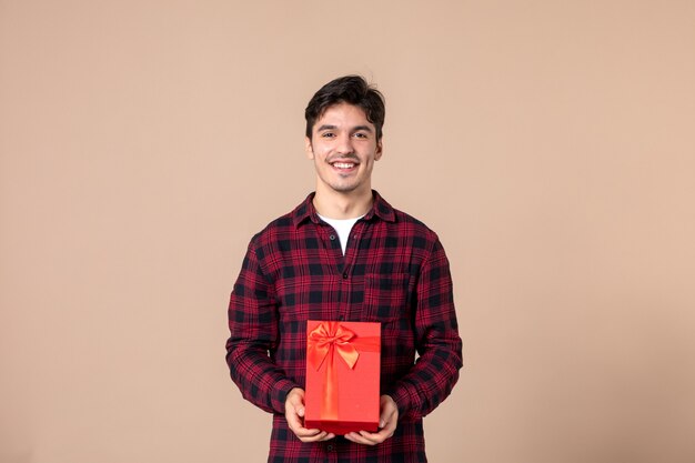 Front view young man holding red package with present for female on brown wall