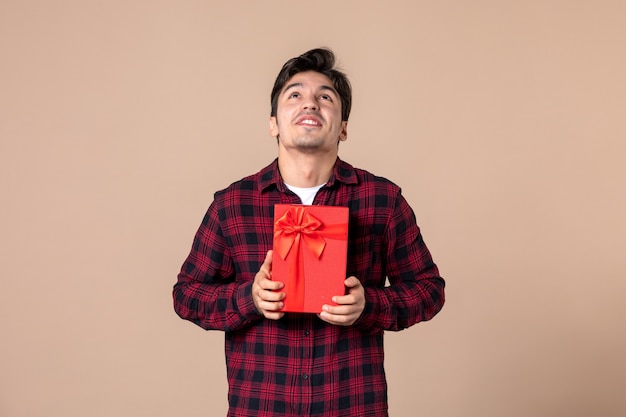 Front view young man holding red package with present for female on brown wall