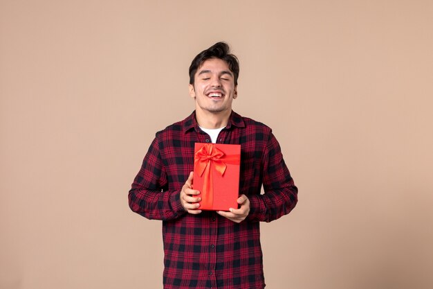 Front view young man holding red package with present for female on brown wall