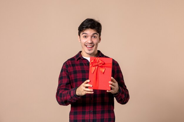 Вид спереди молодой человек, держащий красный пакет с подарком для женщины на коричневой стене
