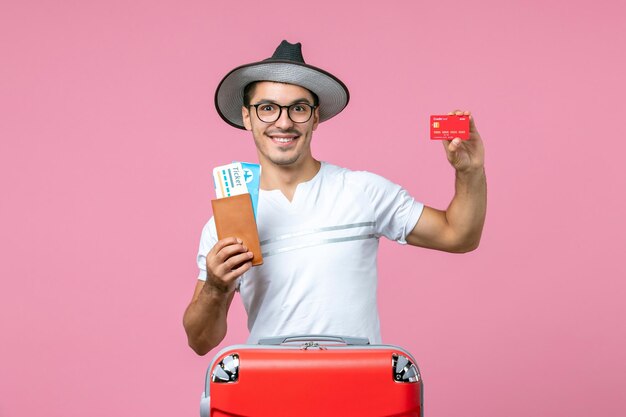 Front view of young man holding tickets and bank card on pink floor summer voyage man plane emotion trip