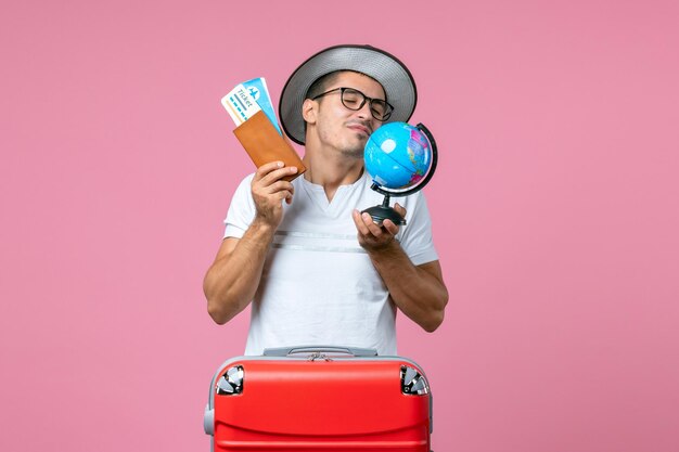 Front view of young man holding tickets and little globe on pink wall