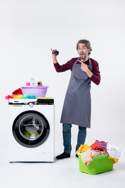 Free photo front view young man holding up card standing near washing machine laundry basket on white wall