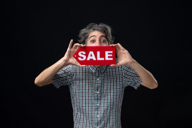 Free photo front view of young man holding up sale sign in front of his mouth standing on dark wall