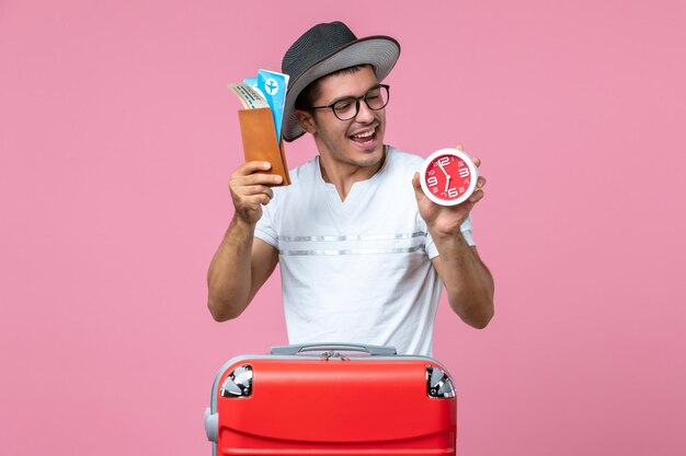 Front view of young man holding vacation tickets and clock on pink wall