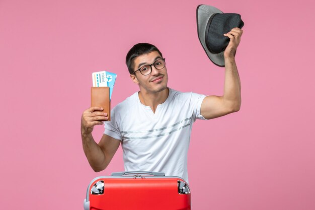 Front view of young man holding vacation tickets and his hat on pink wall