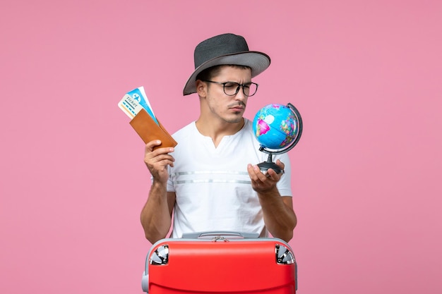 Front view of young man holding vacation tickets and little globe on pink wall