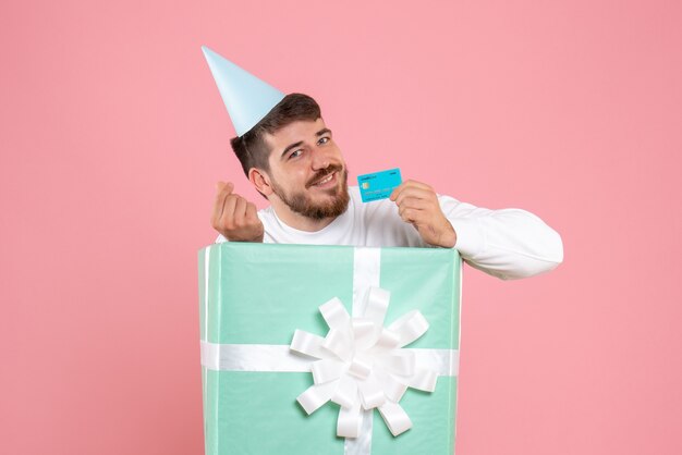 Front view of young man inside present box holding bank card on a pink wall