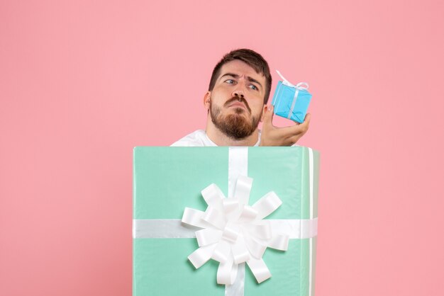 Front view of young man inside present box holding little gift on a pink wall