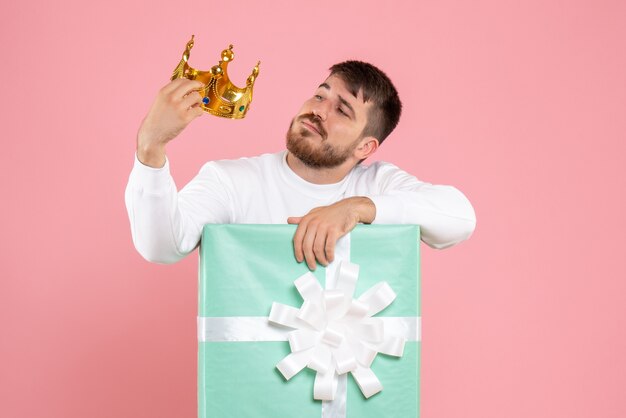 Front view of young man inside present box with crown on his head on pink wall