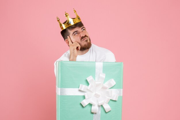 Front view of young man inside present box with crown thinking on pink wall