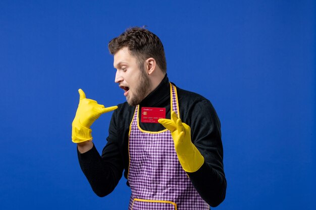 Front view of young man making call me phone gesture holding card on blue wall