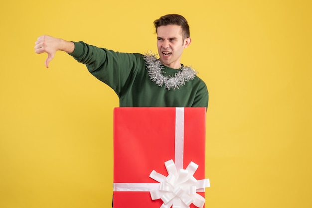 Front view young man making thumb down sign standing behind big giftbox on yellow 