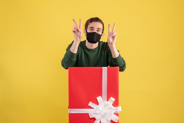 Front view young man making victory sign standing behind big giftbox on yellow 