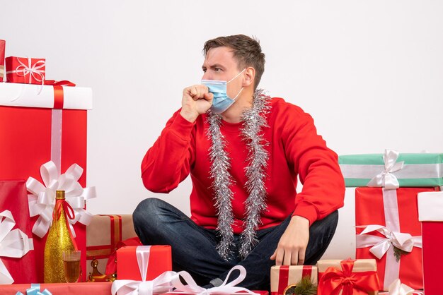 Front view of young man in mask sitting around xmas presents coughing on white wall