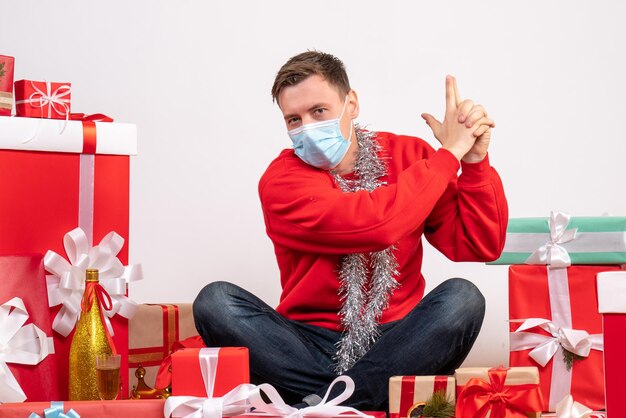 Front view of young man in mask sitting around xmas presents on a white wall