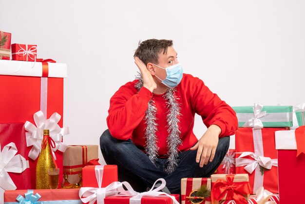 Front view of young man in mask sitting around xmas presents on a white wall