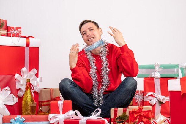 Vista frontale del giovane in maschera seduto intorno ai regali di natale sul muro bianco