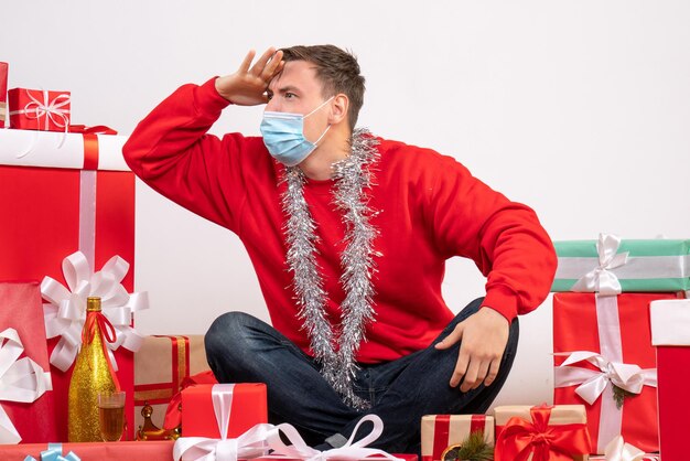 Front view of young man in mask sitting around xmas presents on white wall