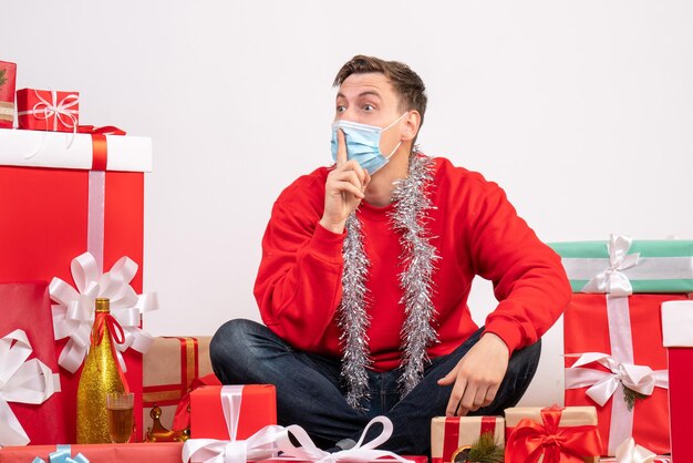 Front view of young man in mask sitting around xmas presents on white wall