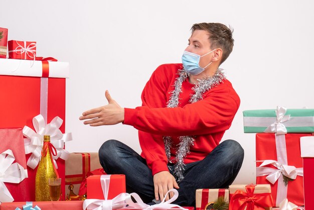 Front view of young man in mask sitting around xmas presents on white wall